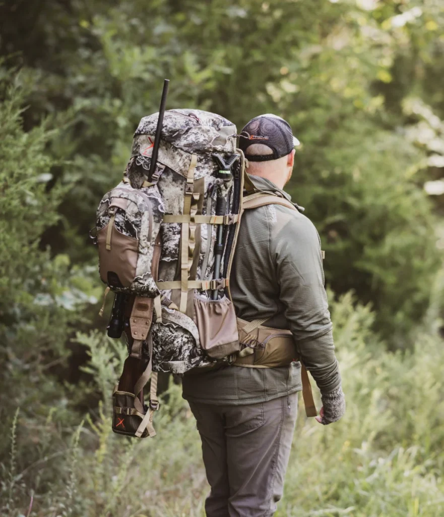 Person hiking through the woods with the ALPS Commander Freighter Frame loaded with camouflage gear, showing the pack’s heavy-hauling external-frame design in the field.