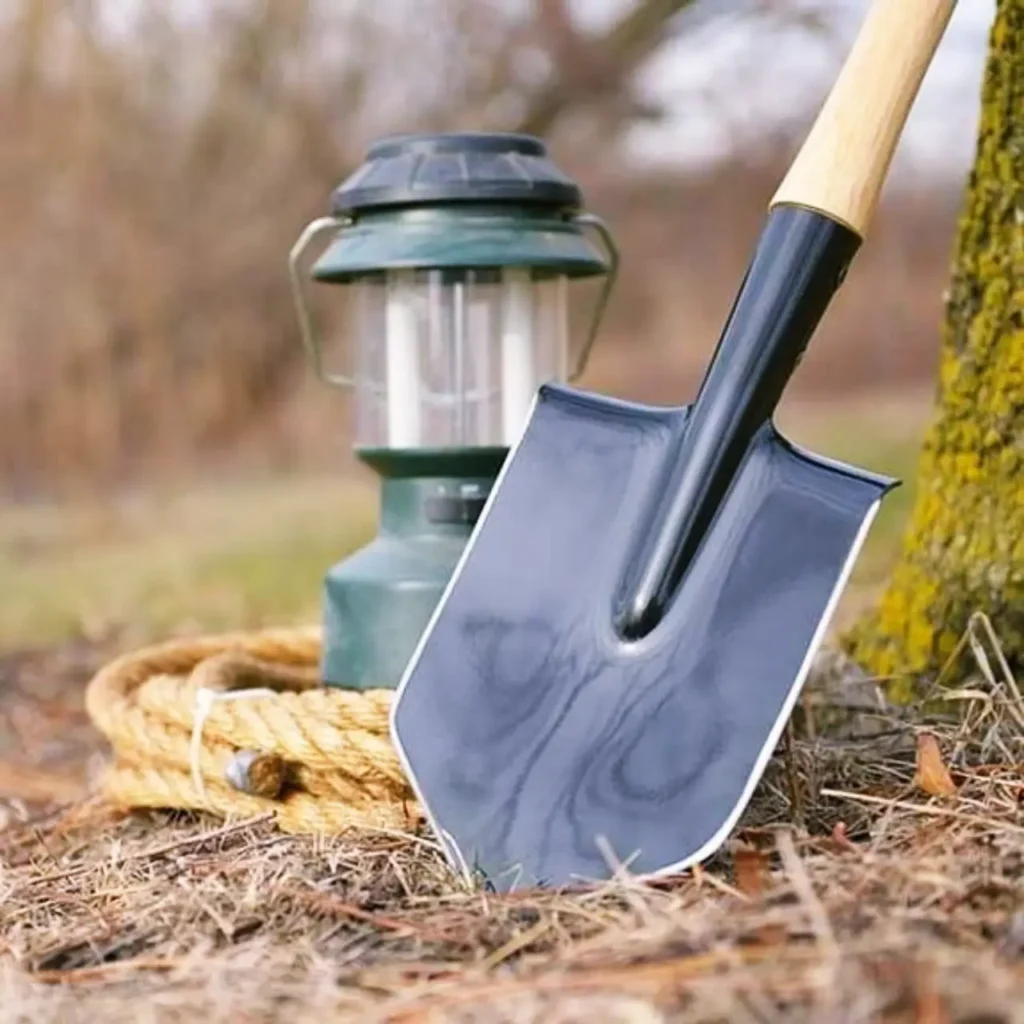 Cold Steel Spetsnaz Shovel set on the ground beside a camp lantern in an outdoor setting, showing its compact steel head and wooden handle.