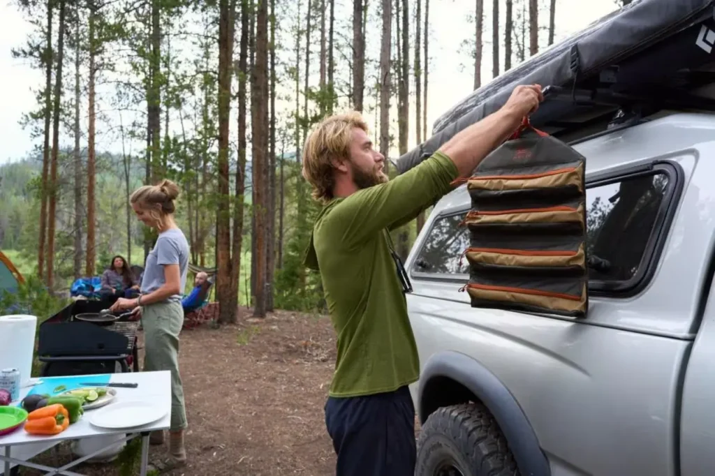 Kelty Chef Roll Bag hanging beside a vehicle during camp kitchen setup at a forest campsite