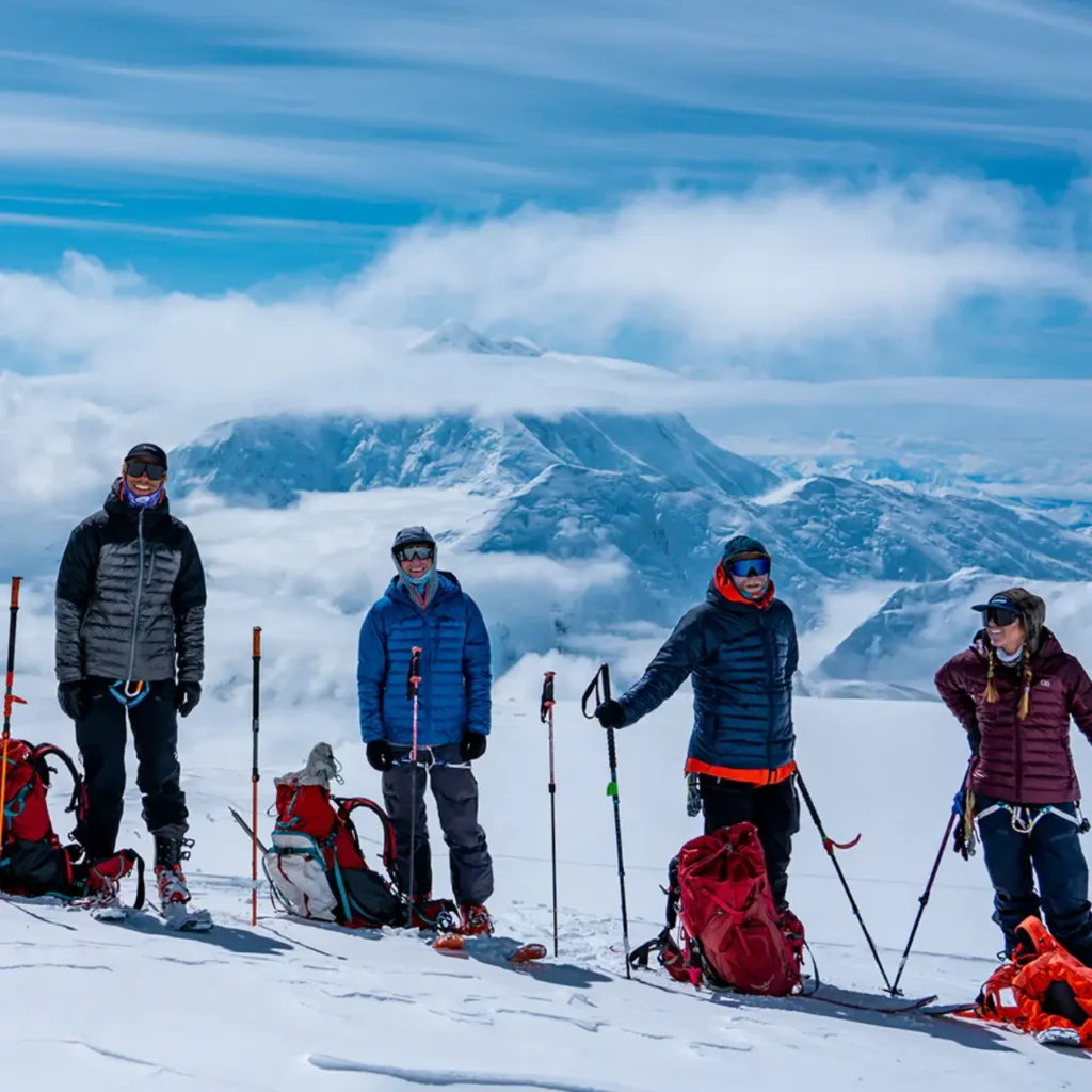 Climbers standing on a snowy alpine ridge wearing the Outdoor Research Helium Down Jacket with glacier-covered mountains in the background.