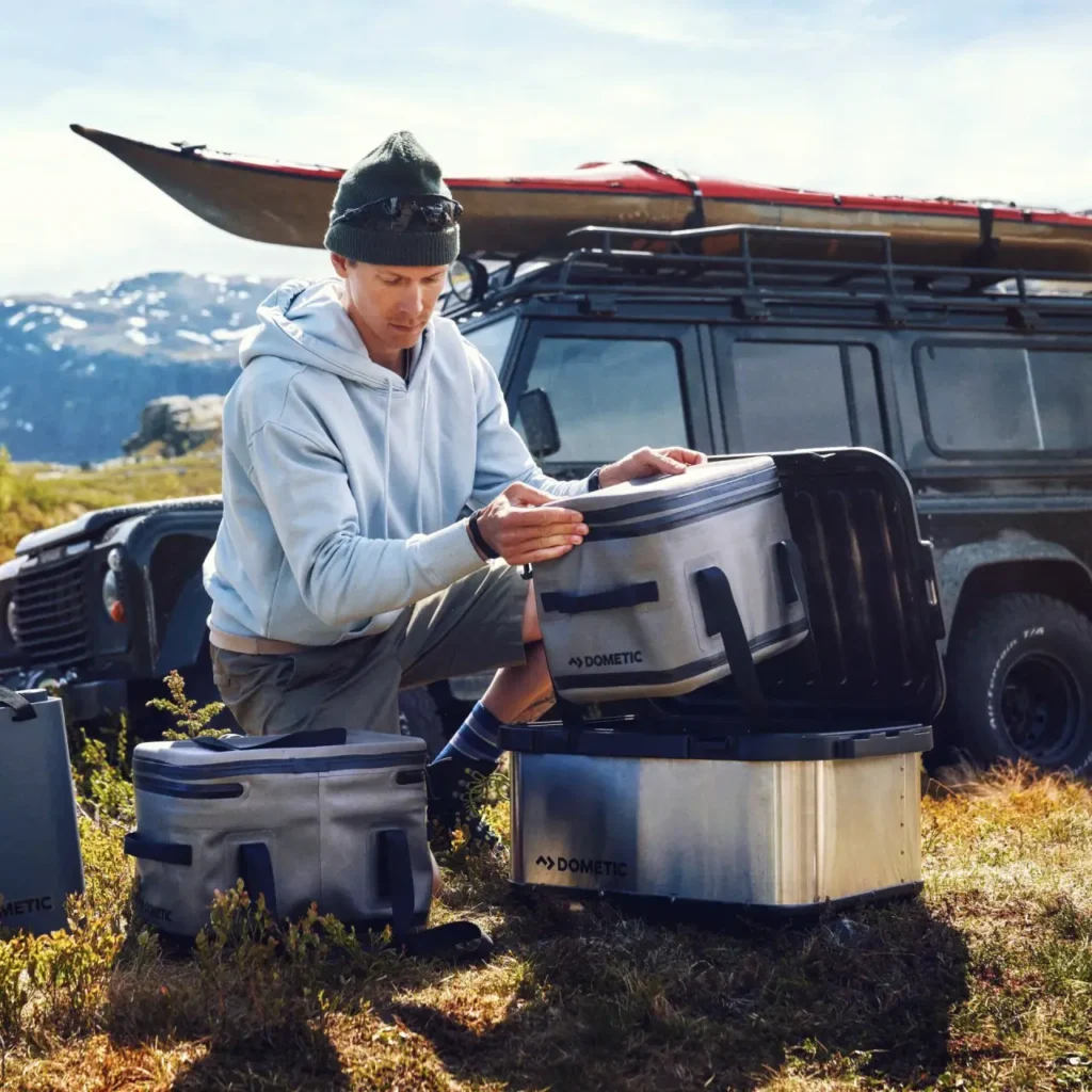 Camper organizing Dometic storage bins beside an off-road vehicle with rooftop kayak in a mountain campsite setting.