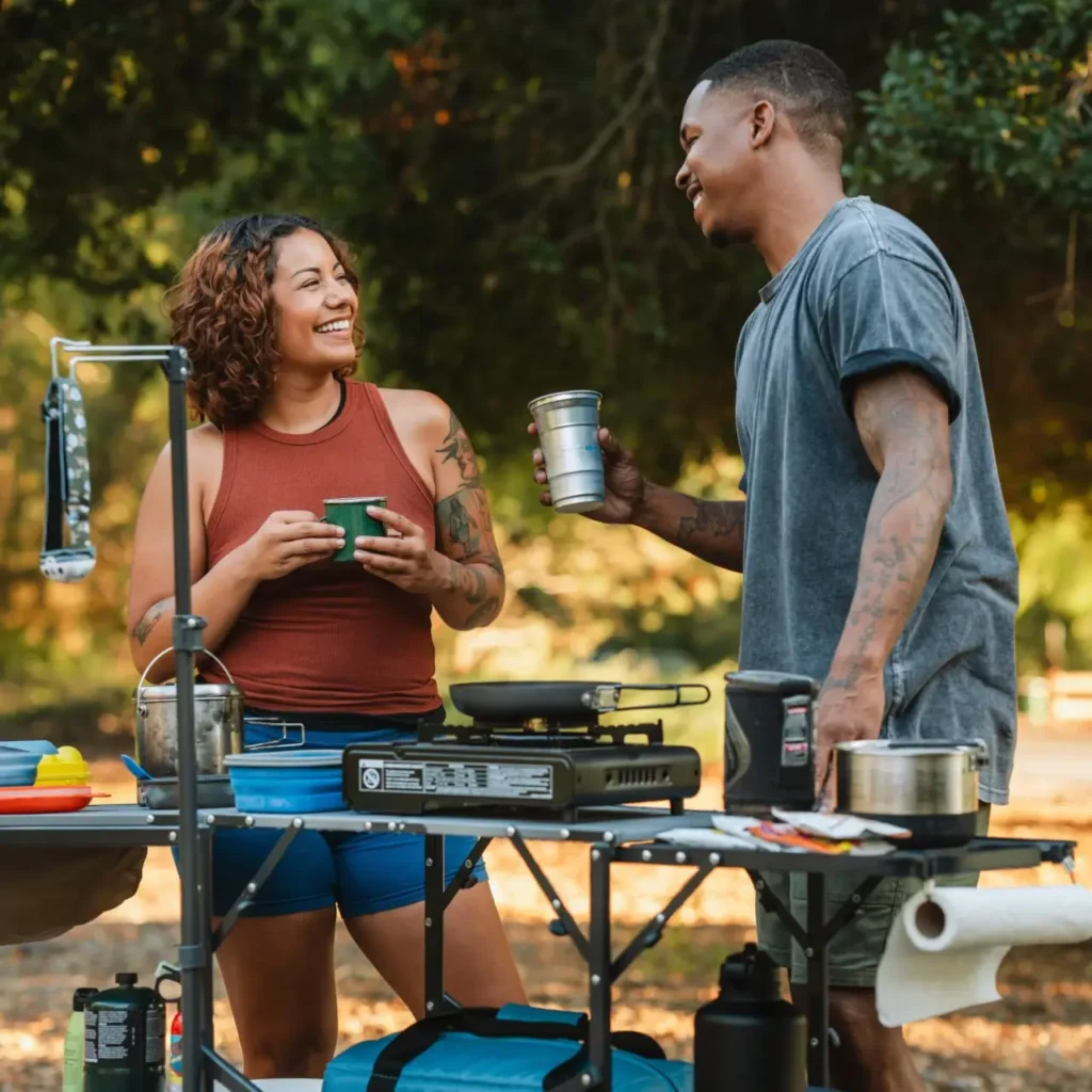 Two campers using one of the best camp kitchen tables with a stove, cookware, mugs, and camp cooking gear at a sunny campsite.