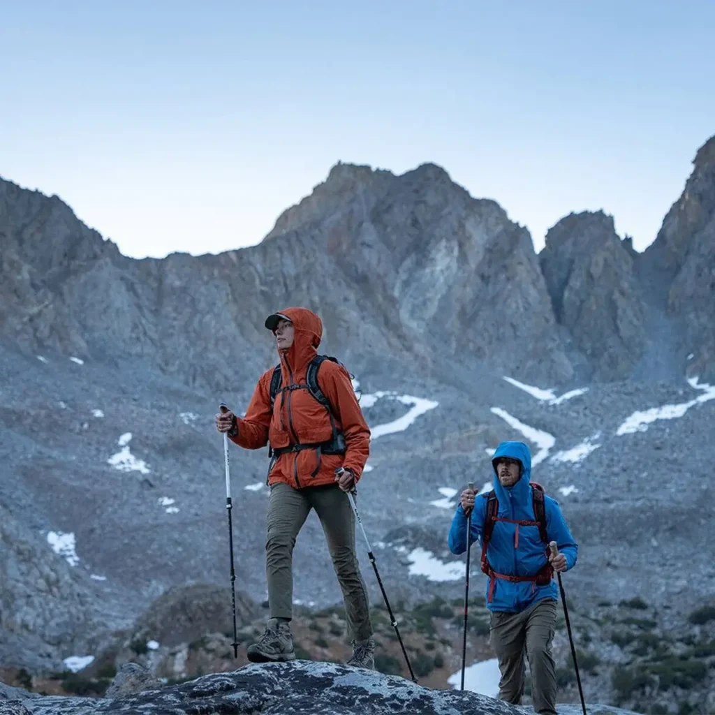 Two hikers wearing Osprey Talon 22 and Osprey Tempest 20 daypacks with trekking poles on a rocky alpine trail surrounded by mountain peaks.