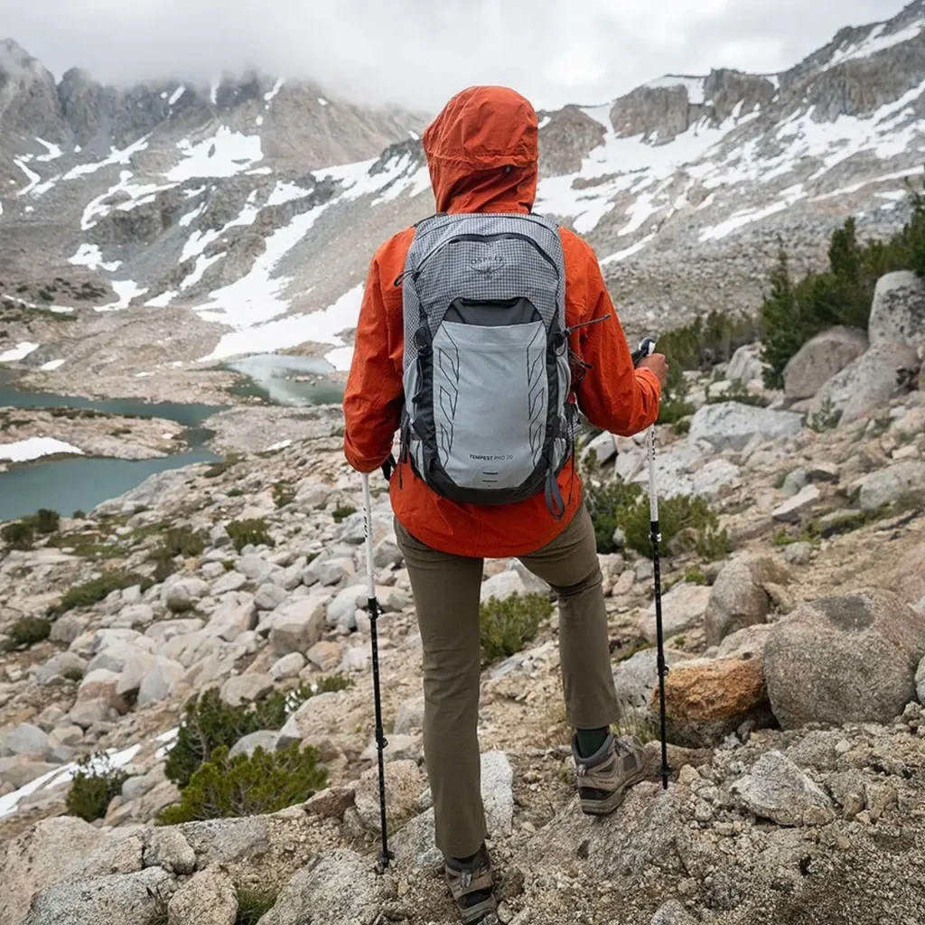 Beginner hiker wearing an Osprey Tempest 20 daypack on a rocky mountain trail, showing hiking essentials for beginners including trekking poles, rain jacket, hiking pants, and trail shoes.