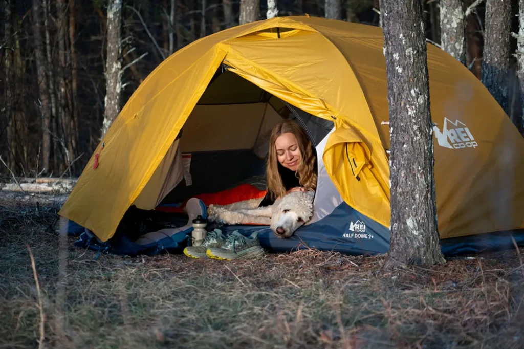 Camping essentials for beginners shown in a cozy REI tent setup at a forest campsite with a sleeping pad, shoes, water bottle, and dog resting at the entrance