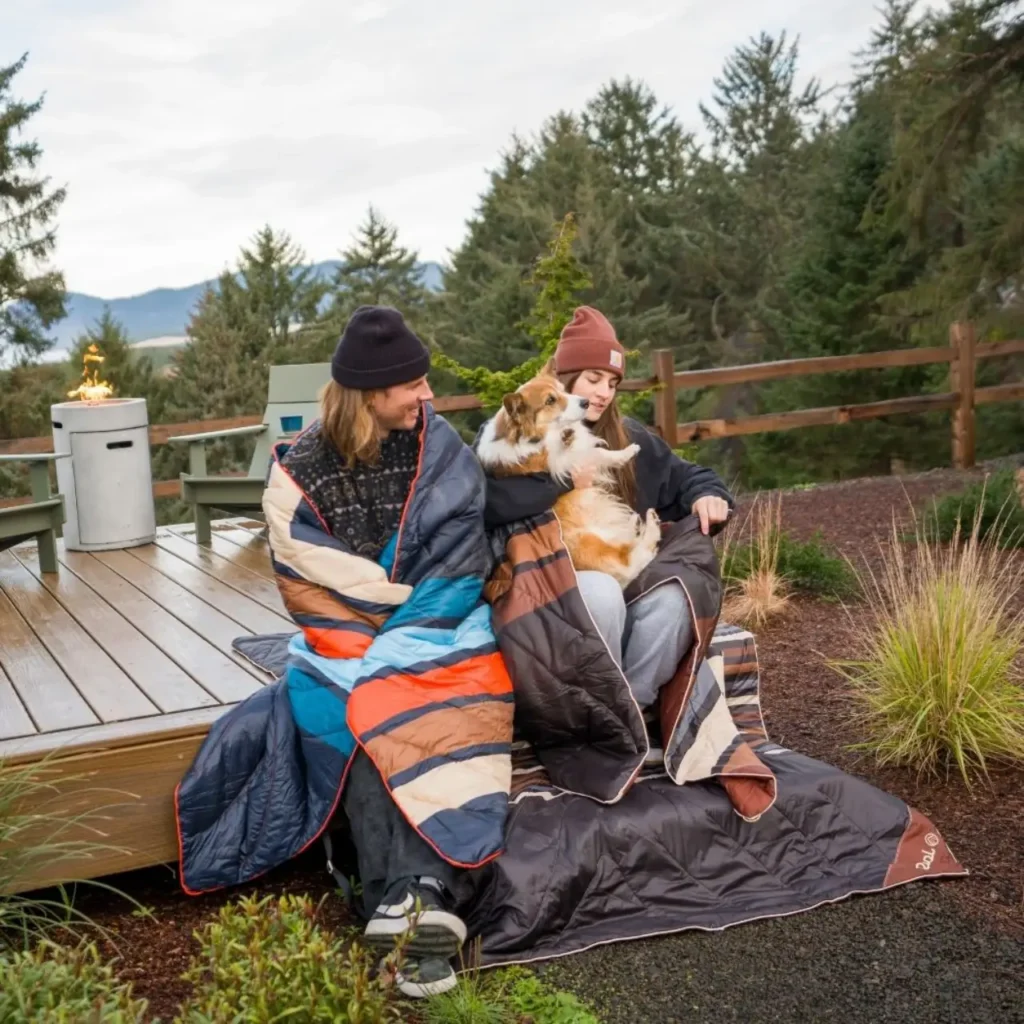 Two campers wrapped in Rumpl Original Puffy Blankets with a dog at a cozy outdoor campsite.