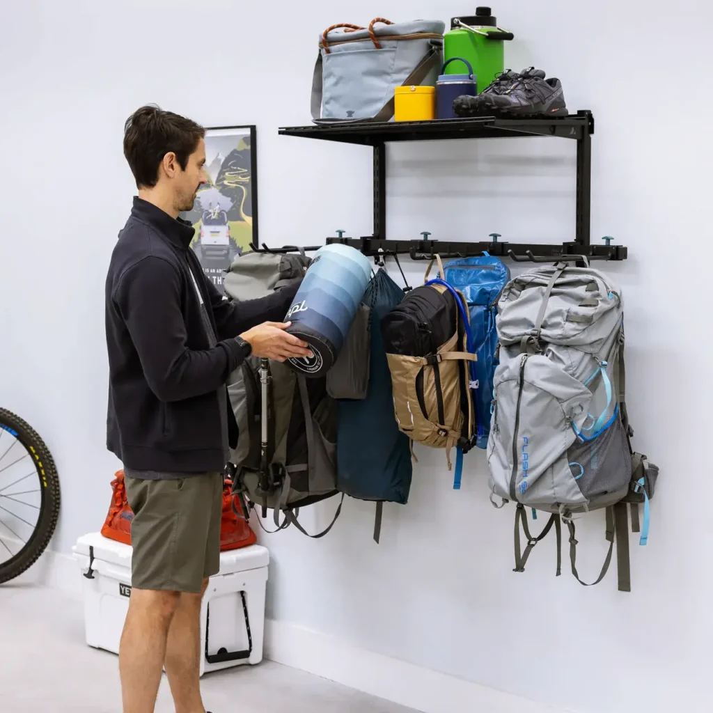 Wall-mounted outdoor gear storage system with hiking backpacks, a rolled sleeping pad, shoes, water bottles, and camping essentials organized neatly in a garage or gear room.