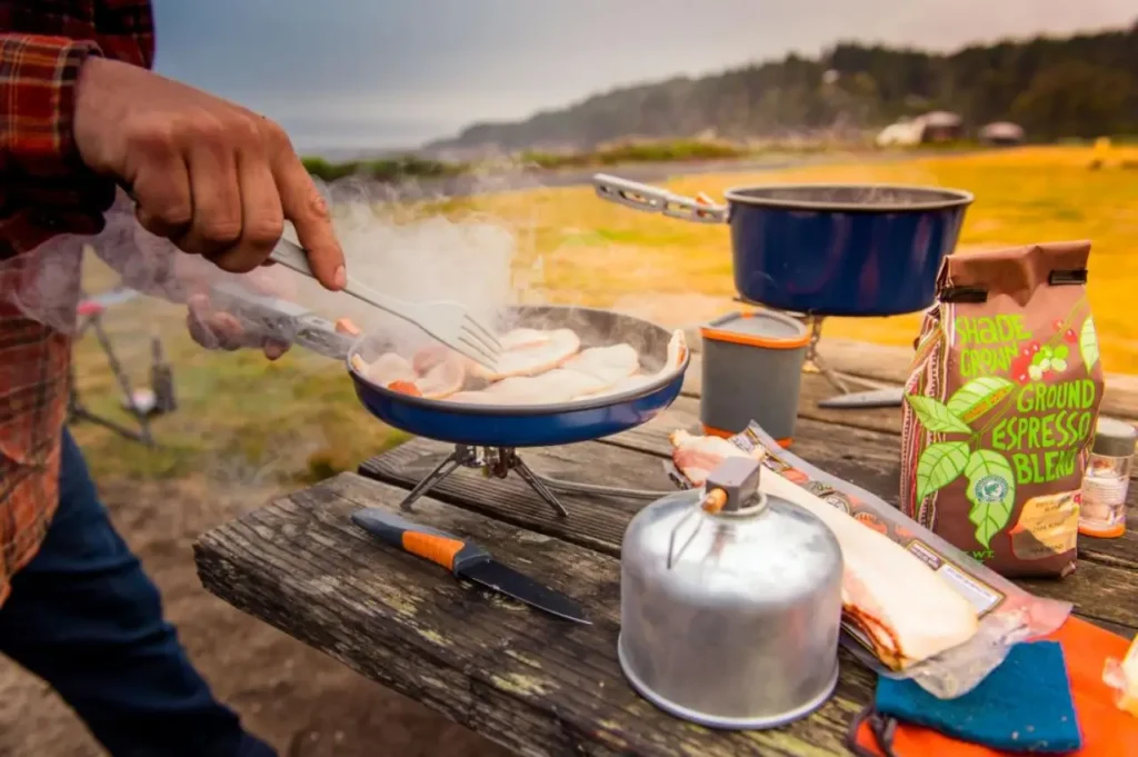 person cooking breakfast on a camp stove at a picnic table with camp kitchen gear and coffee outdoors