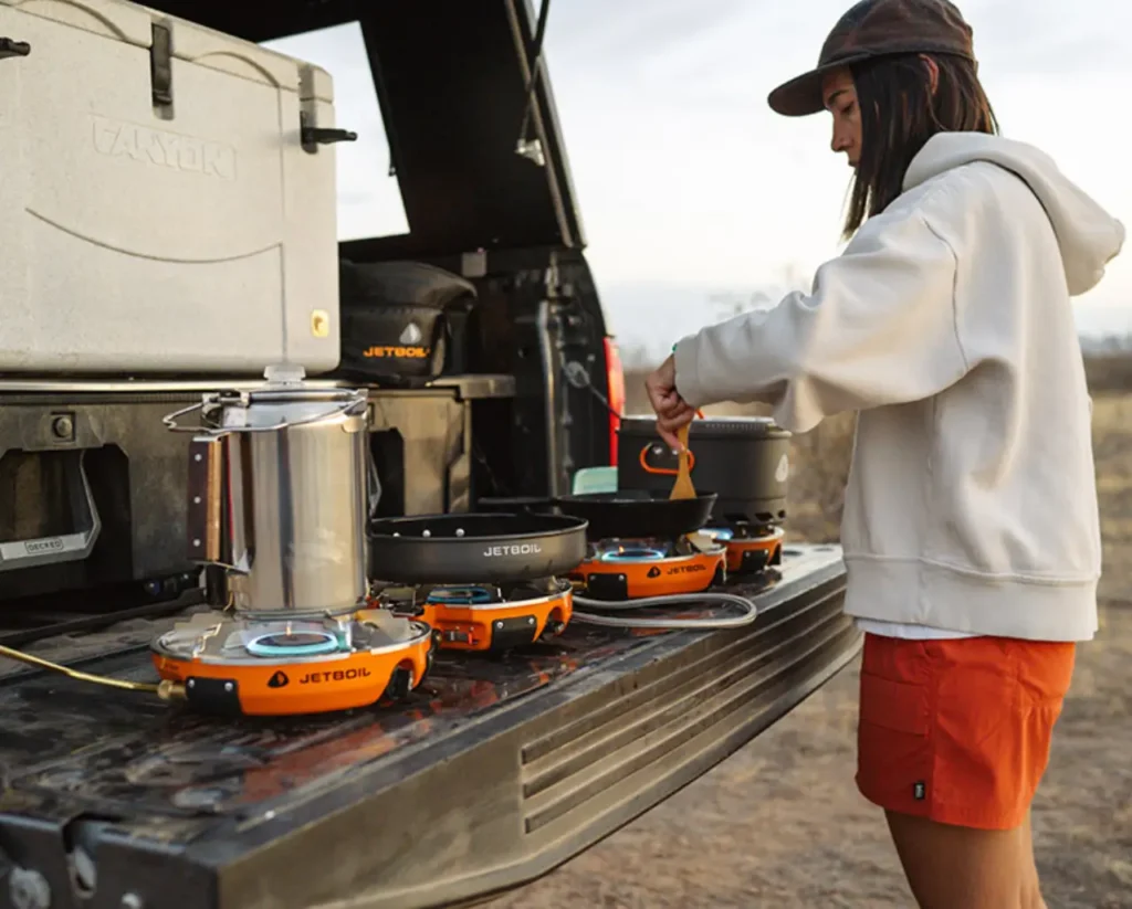 Tailgate car camping kitchen setup showing portable camp stoves and cookware in use, illustrating the best camping stoves for car camping during an outdoor meal setup.