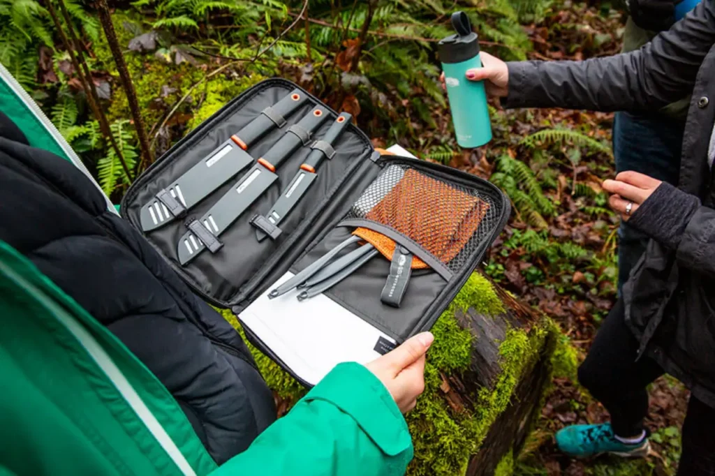 GSI Outdoors Santoku Knife Set opened at a forest campsite with knives, utensils, and a cutting board ready for outdoor meal prep.