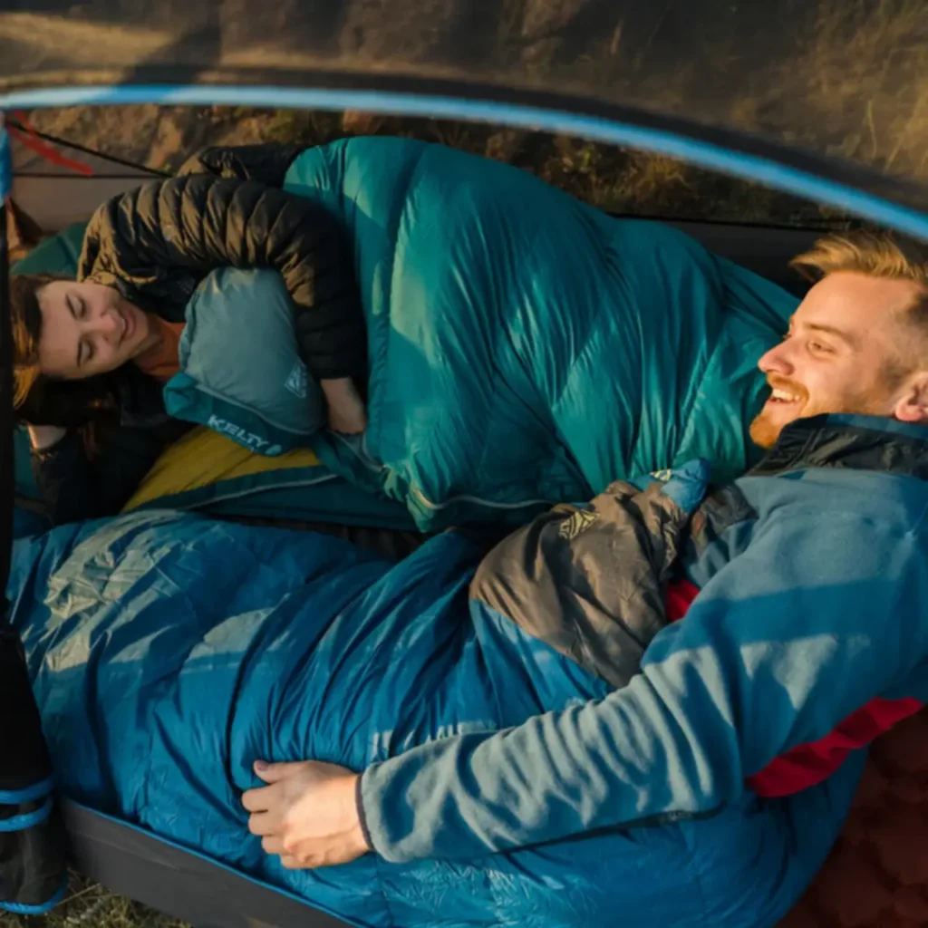 Two campers relaxing inside a tent with the Kelty Cosmic 20 sleeping bag during a beginner-friendly weekend camping trip.