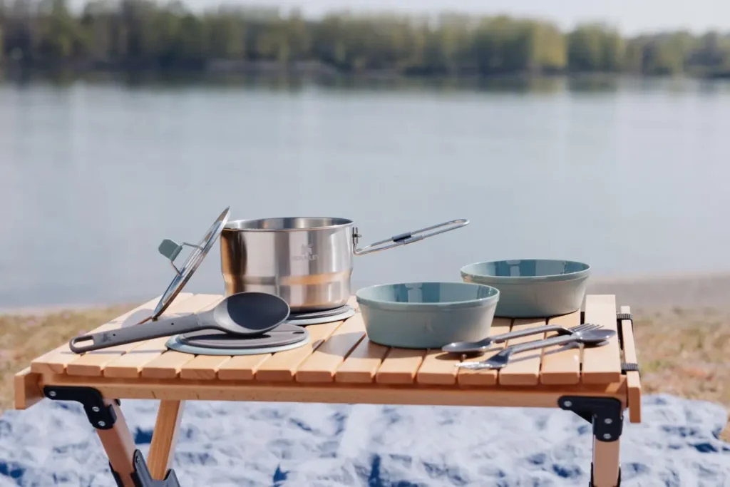 Best camp cookware sets displayed on a folding camp table beside a lake, with a stainless steel pot, bowls, utensils, and outdoor cooking accessories.