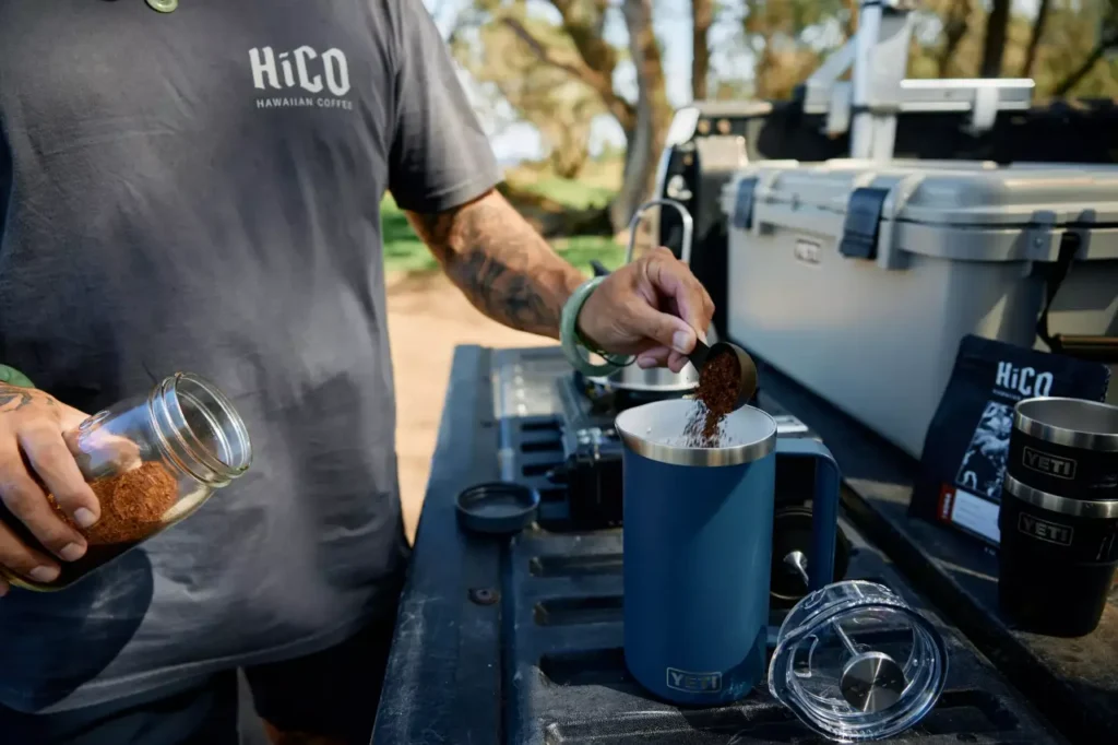 YETI Rambler French Press being filled with ground coffee at an outdoor camp kitchen setup.