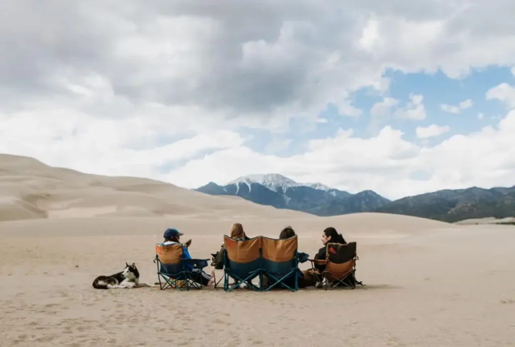 Kelty Low Loveseat set up on sandy desert terrain with campers relaxing and mountain views in the background.