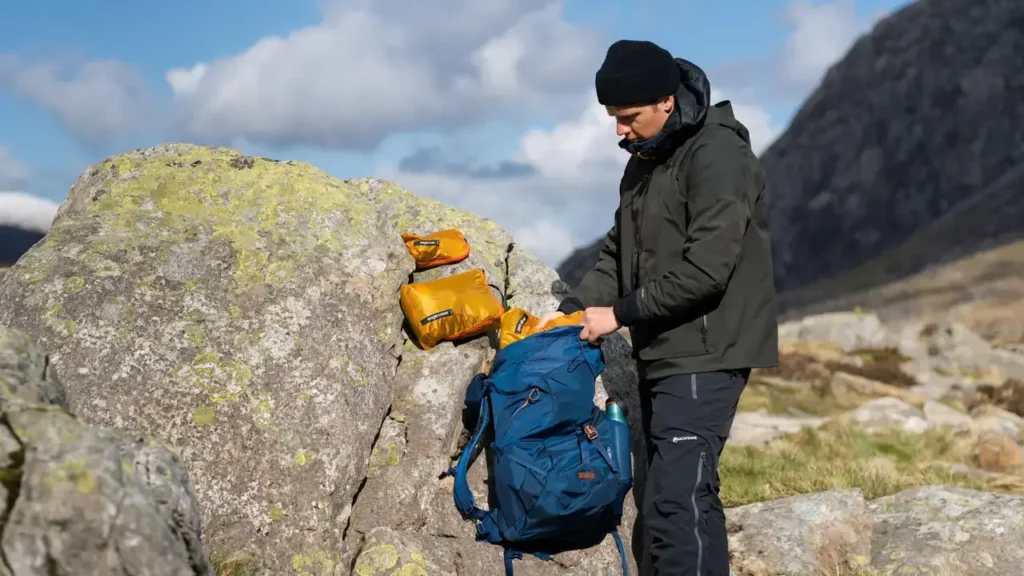 Hiker organizing Eagle Creek Pack-It Isolate packing cubes beside a blue backpack in a rocky mountain landscape.