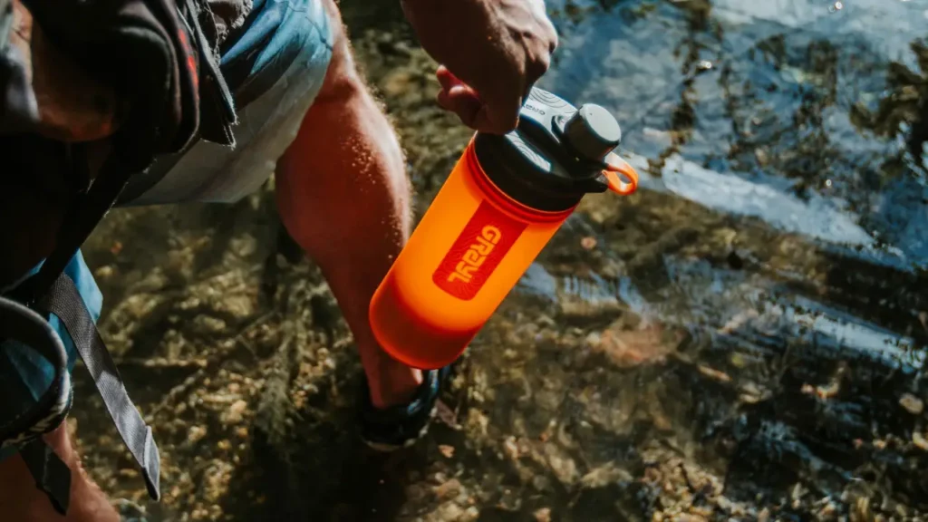 Hiker holding an orange Grayl GeoPress Water Purifier 24 oz. bottle near a clear stream during an outdoor hike.
