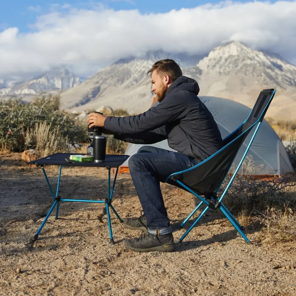 Camper using a Helinox Table One for coffee and small essentials beside a tent at a mountain campsite.