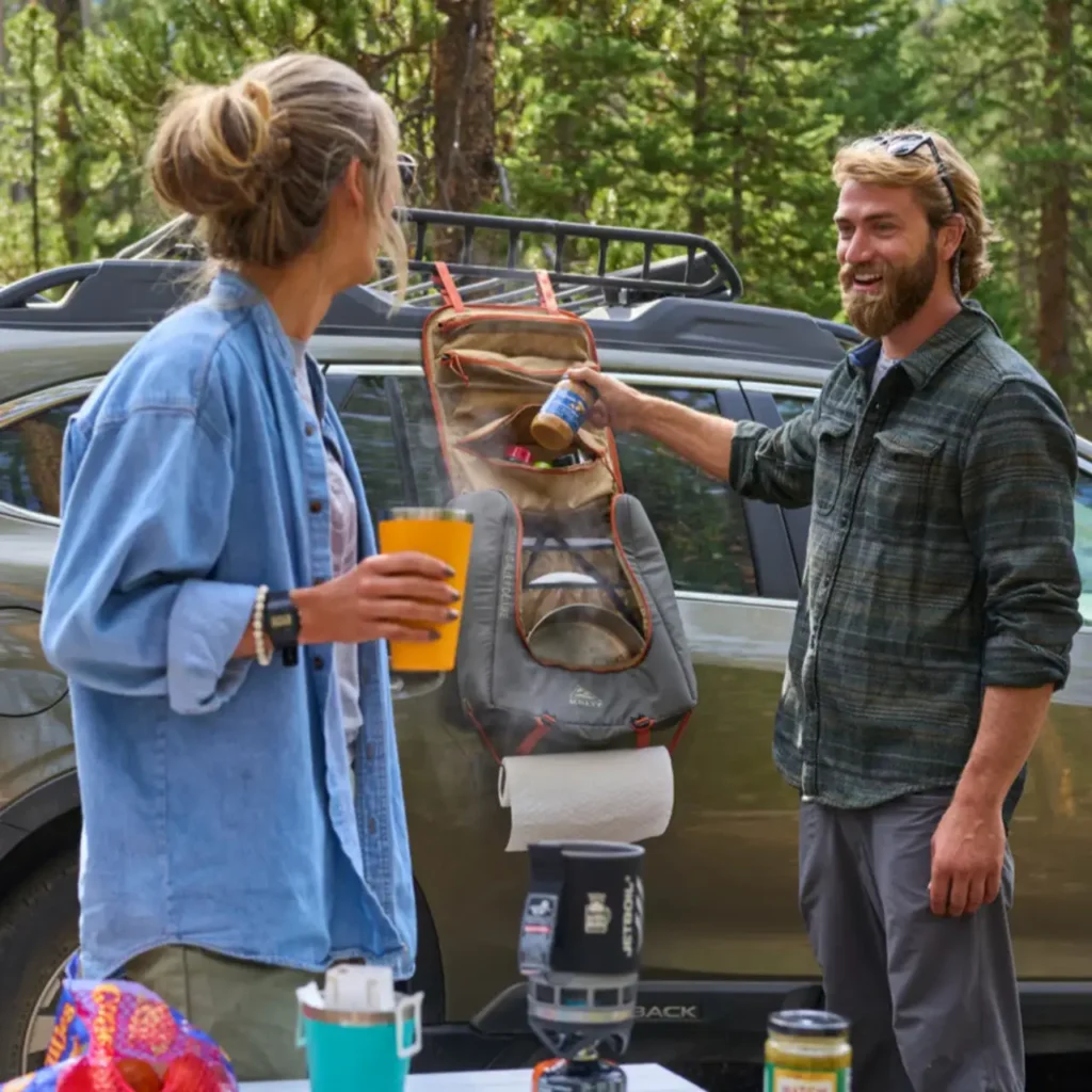 Kelty Camp Galley Deluxe Bag hanging from the side of a vehicle at a forest campsite, with campers organizing spices, coffee gear, and paper towels.