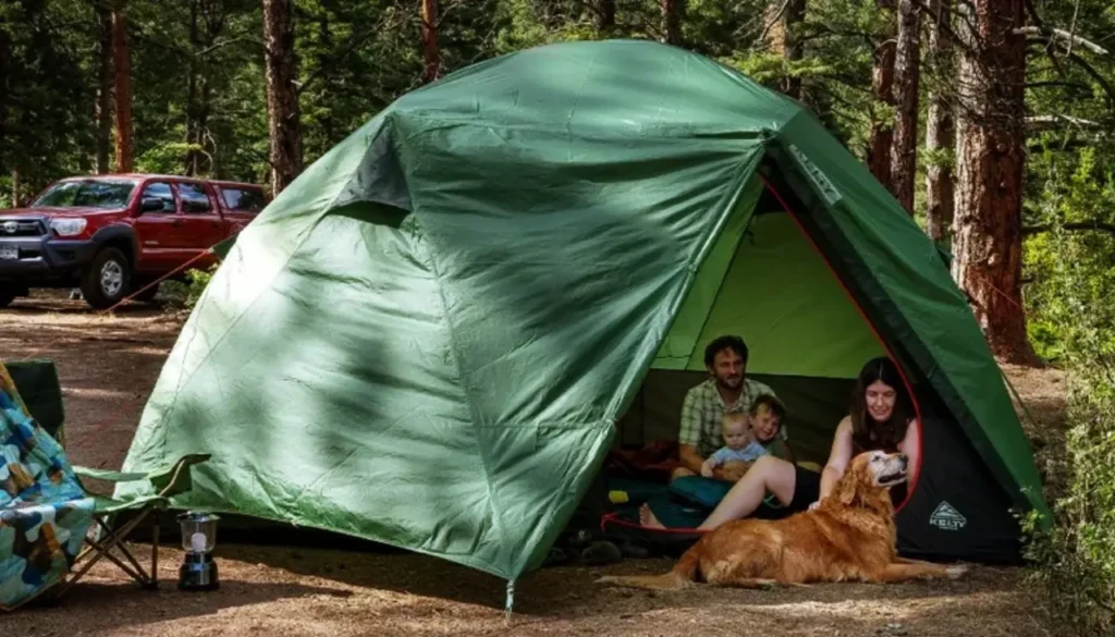 Family relaxing inside the spacious Kelty Wireless 8 Tent at a wooded campground with camping chairs, a lantern, and a dog nearby.