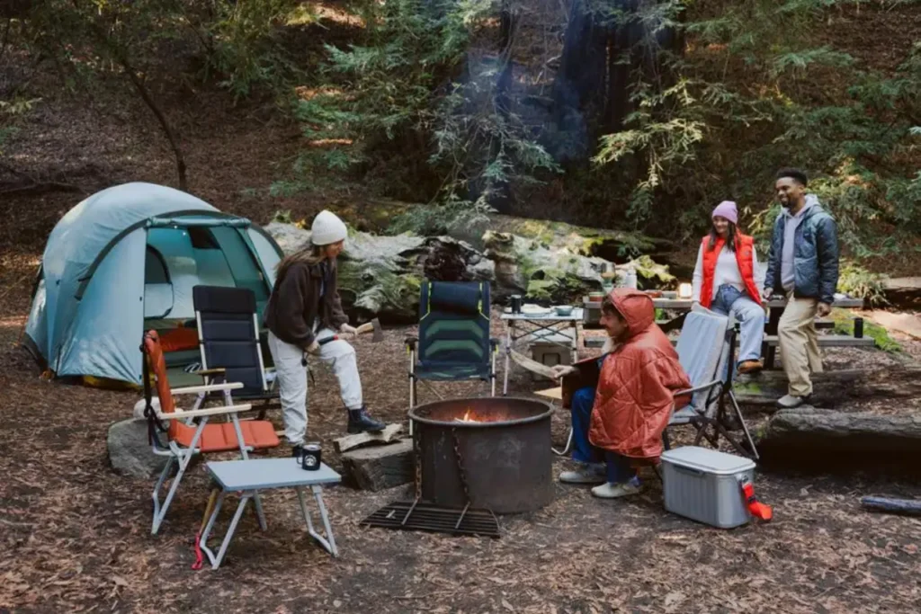 REI Co-op Westward 6 Tent set up at a wooded family campsite with camp chairs, a cooler, a fire ring, and campers preparing for a comfortable weekend outdoors.