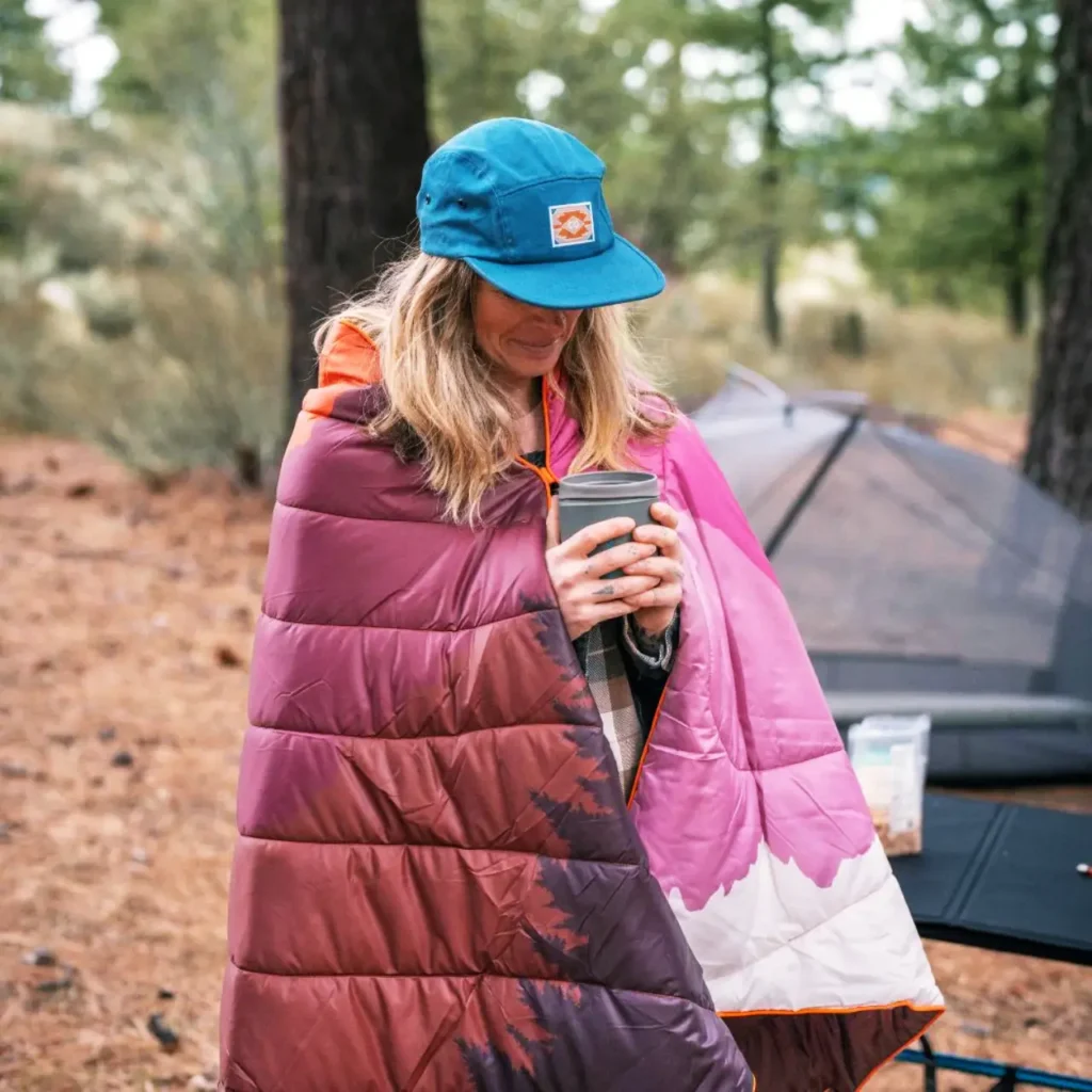 Camper wrapped in a Rumpl Original Puffy Blanket while holding a warm drink beside a tent.