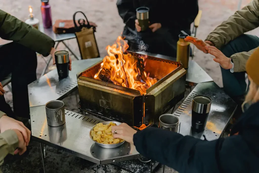 Campers gathered around a Snow Peak Takibi Fire & Grill with a wood fire burning at a campsite.