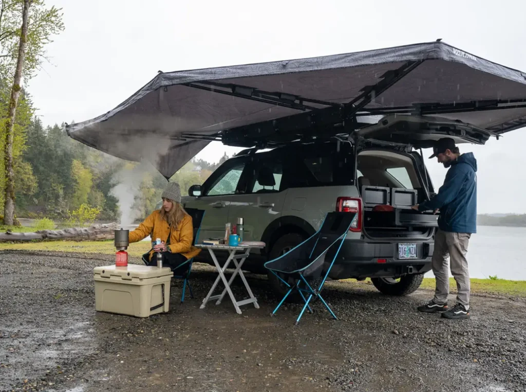 Yakima OverNOut 270 XL Awning deployed over an SUV campsite in the rain, creating covered space for cooking, chairs, and tailgate gear.