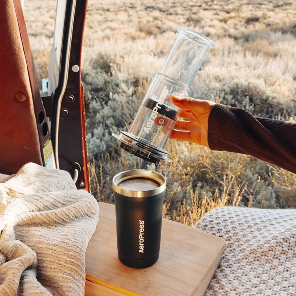 AeroPress Go Travel Coffee Press brewing coffee into an insulated mug beside an open camper van in a desert campsite.