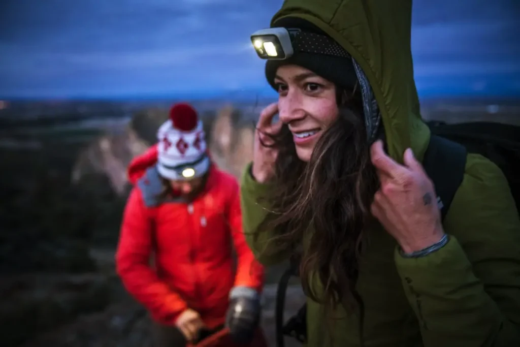 Two hikers wearing camping headlamps on a trail at dusk, including a woman adjusting her hood in the foreground.