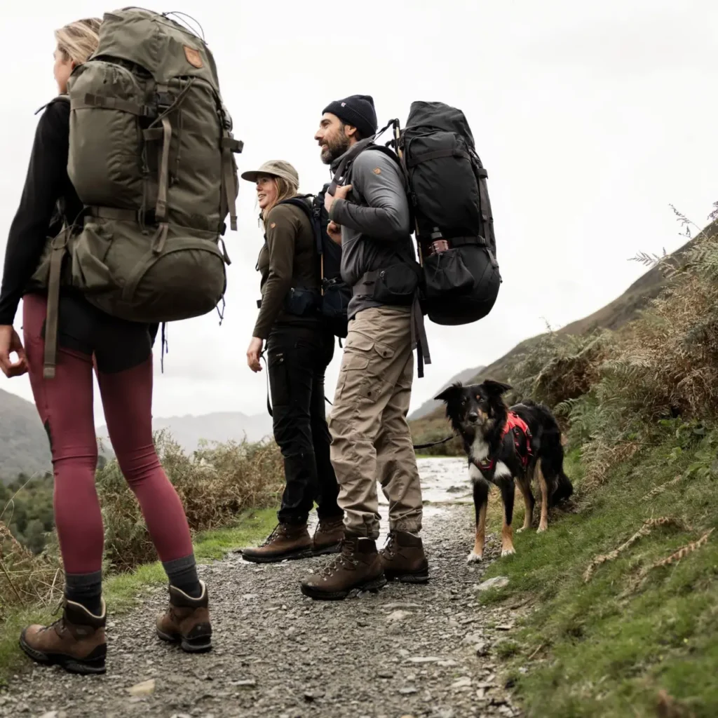 Three hikers wearing different hiking pants and carrying large backpacks on a mountain trail with a dog.