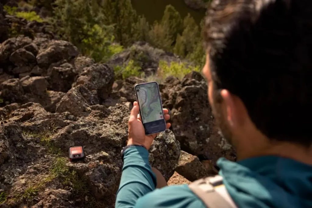 Hiker using a smartphone for GPS navigation beside a Garmin inReach Messenger Plus on a remote rocky trail.