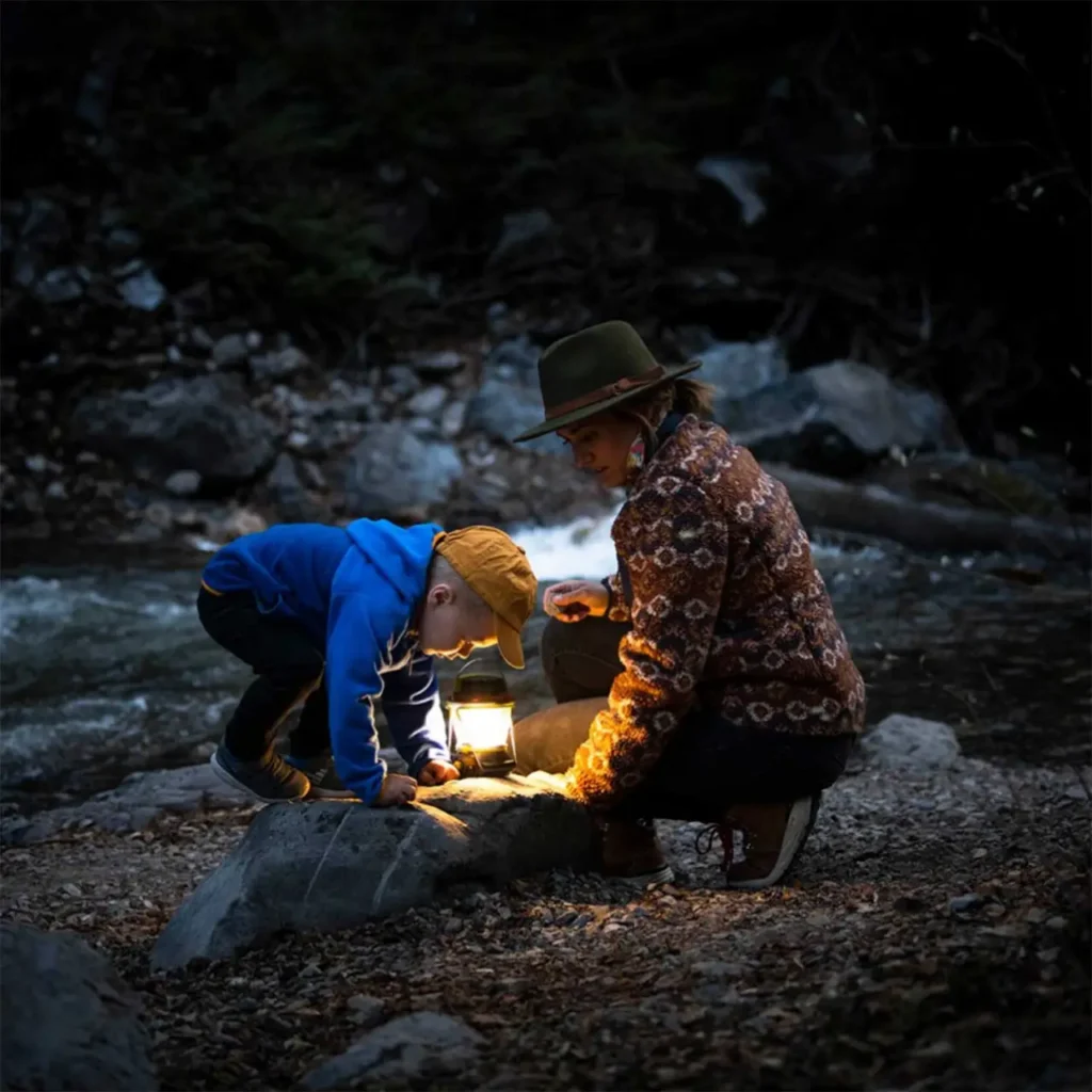 Child and adult using a glowing Goal Zero Lighthouse 600 camping lantern beside a creek at dusk.