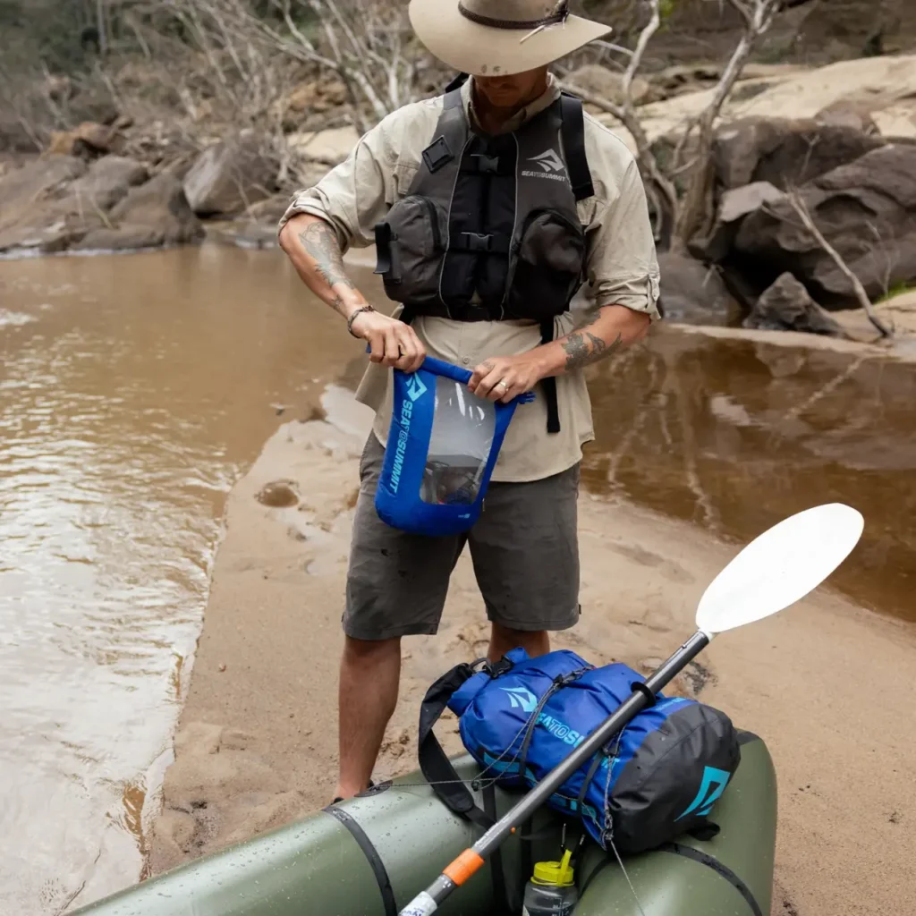 Paddler closing a blue Sea to Summit dry bag beside a loaded packraft on a rocky riverbank.