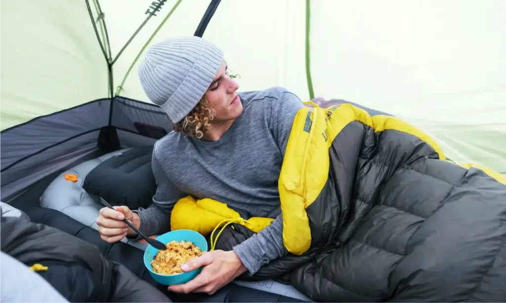 Camper eating from a blue bowl with a long-handled camping spoon while resting in a sleeping bag inside a tent.