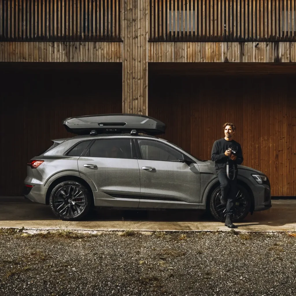 Gray SUV with a Thule Motion 3 L roof cargo box parked beside a wooden building, with a traveler standing near the front of the vehicle.