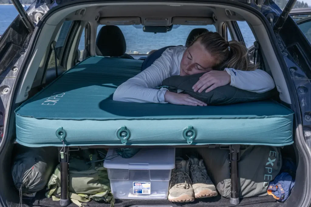 Camper resting on an EXPED MegaMat Duo car camping mattress set up inside the back of an SUV beside a lake.