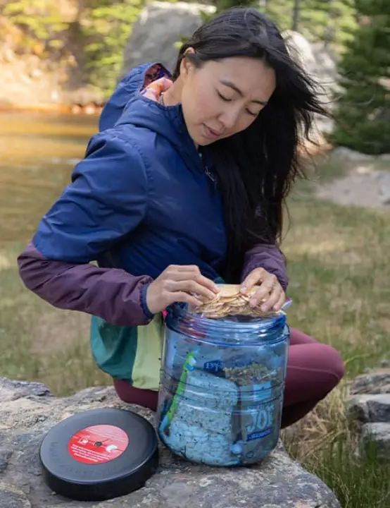 Backpacker packing food into a BearVault BV475 Trek bear canister beside a mountain lake.