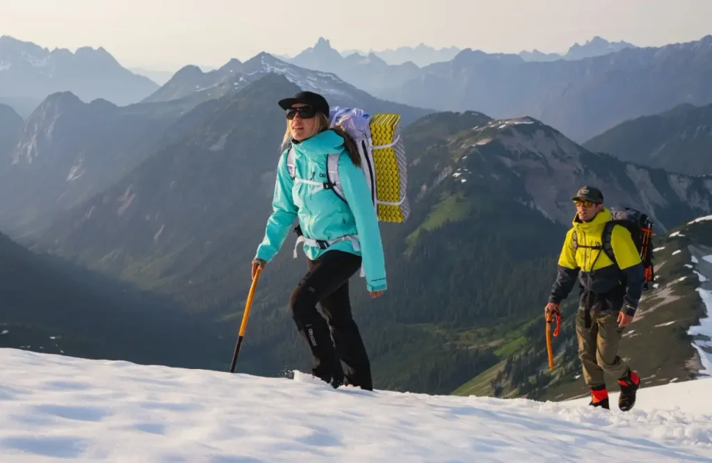 Two hikers wearing cold weather hiking gear and carrying backpacks while climbing a snowy mountain trail.