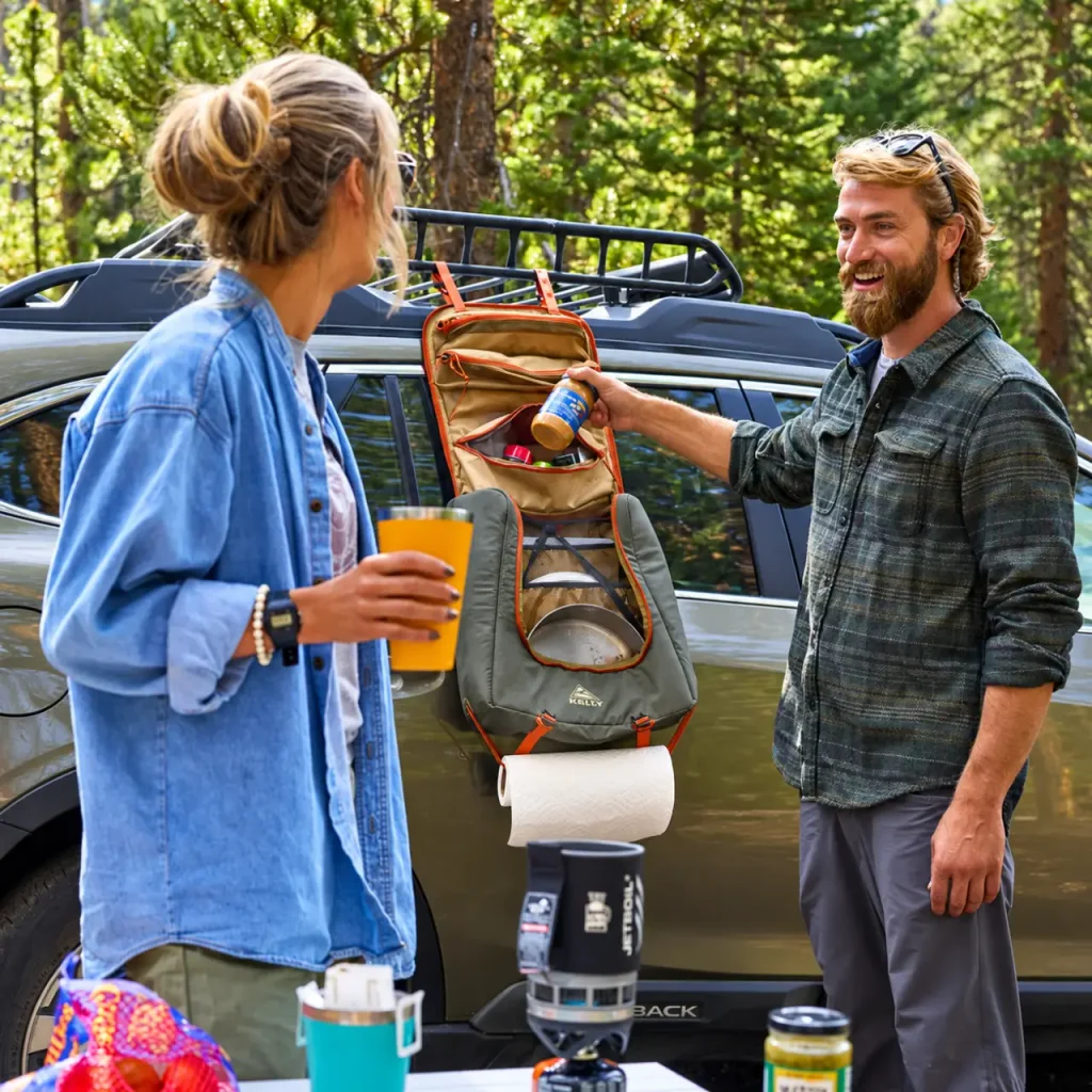 Kelty Camp Galley Deluxe hanging organizer with cooking utensils, cookware, and kitchen accessories beside an SUV at a forest campsite, helping reduce camping cookware rattling during travel.