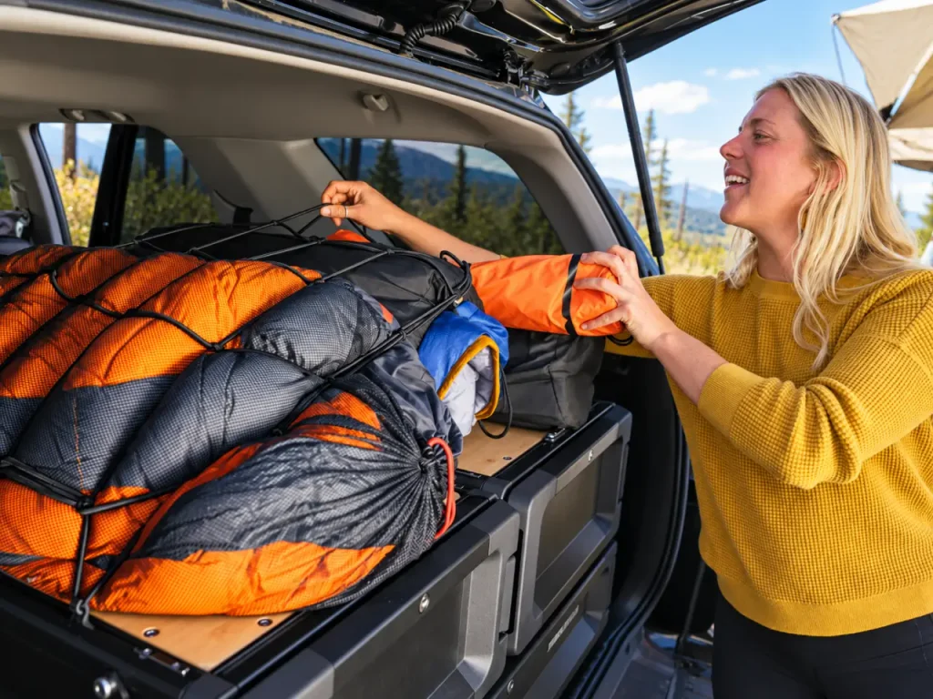 Woman securing sleeping bags and camping gear with a Yakima MOD Stretch Net over storage drawers in the rear cargo area of an SUV.